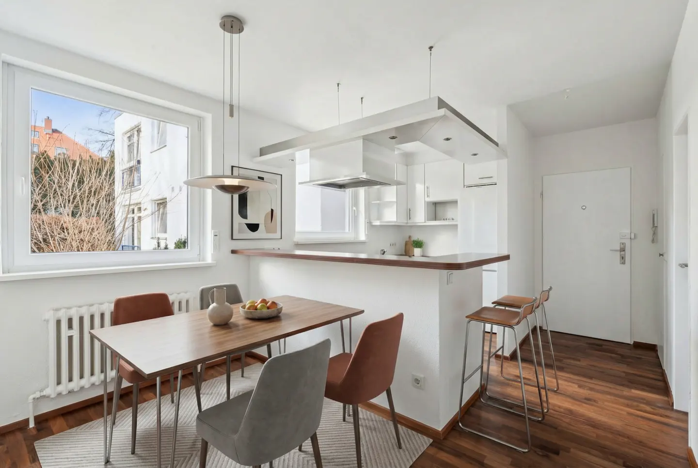 Bright, modern apartment interior with a wood dining table, gray and brown chairs, and a white kitchen with bar stools. A large window overlooks a building.