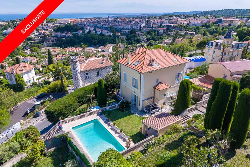 Aerial view of a yellow two-story house with a red tile roof and a blue pool in the yard. A red banner reads "Exclusivite."
