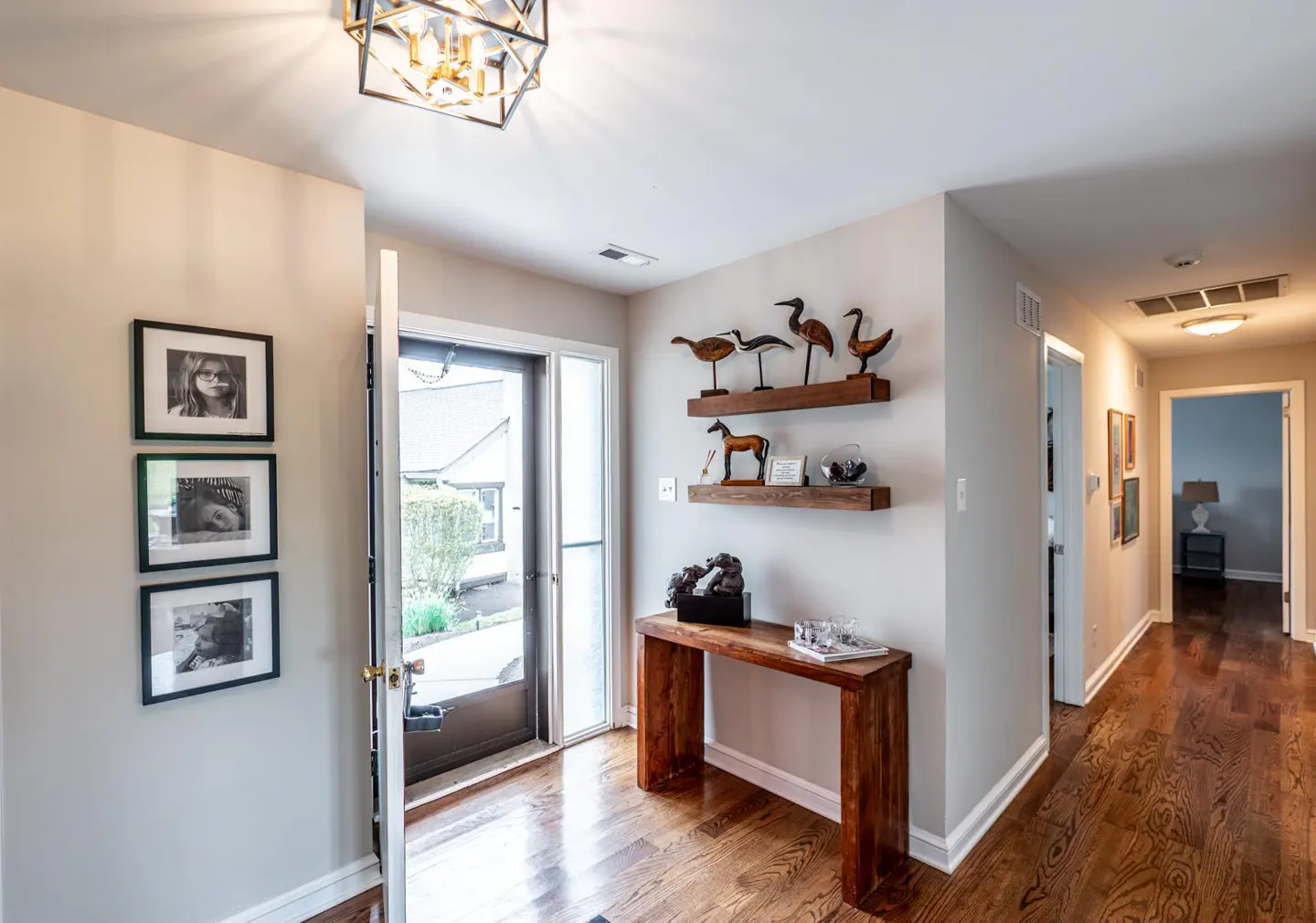 Entryway with wood floors, beige walls, and an open front door. A wood table sits below shelves with bird and horse figurines. Black and white photos hang on the left wall.