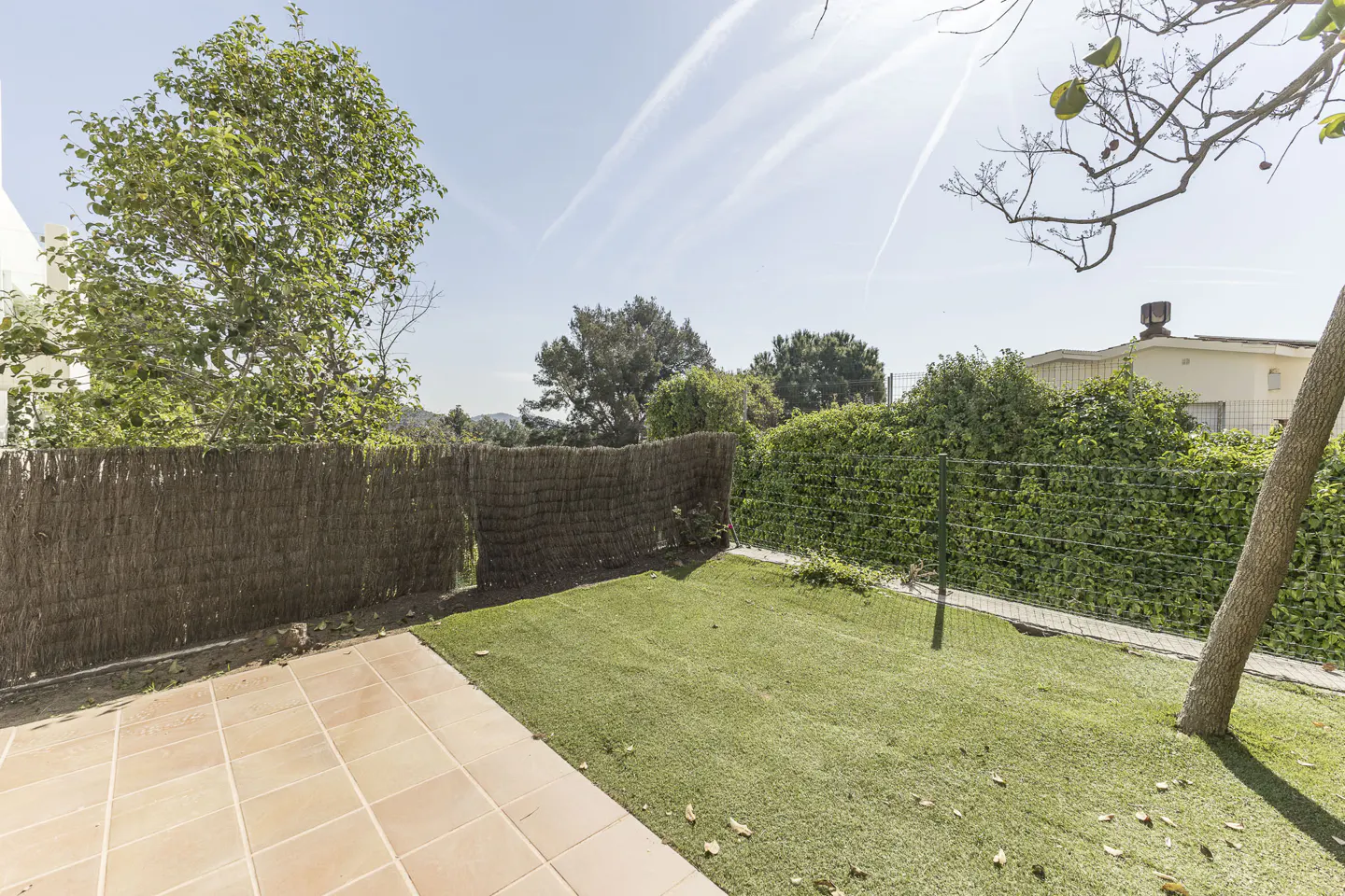 A backyard with brown tile, green grass, and a tall brown fence under a blue sky.