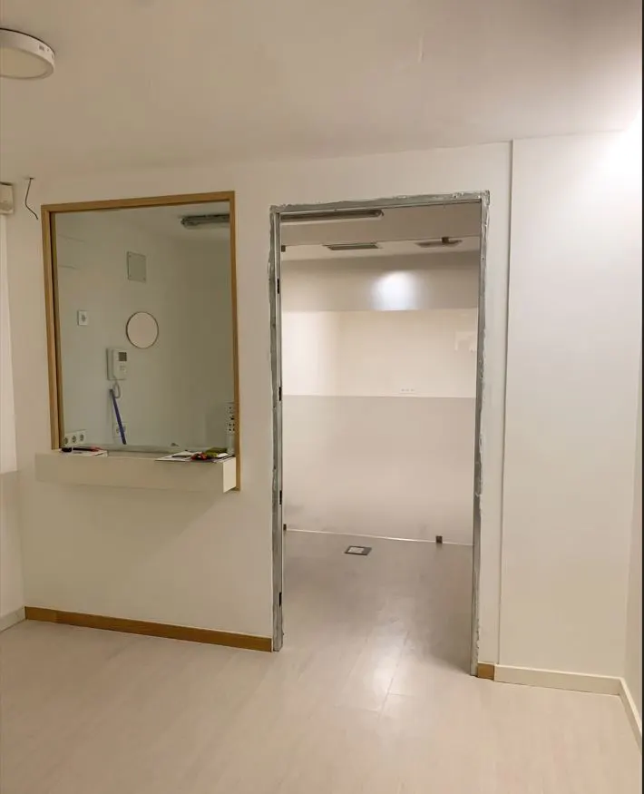 A bright, empty room with white walls and light flooring. A window with a counter and an open doorway are visible.