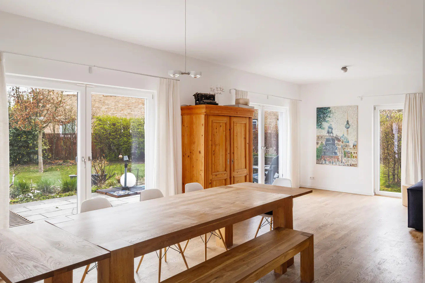 Bright dining room with a long wooden table and bench, white walls, and large windows overlooking a green garden. A wooden cabinet stands against the wall.