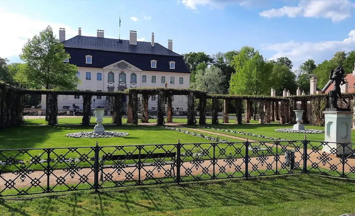 Exterior view of a large, white mansion with a black roof, seen from a green lawn with a black iron fence and vine-covered pergola.