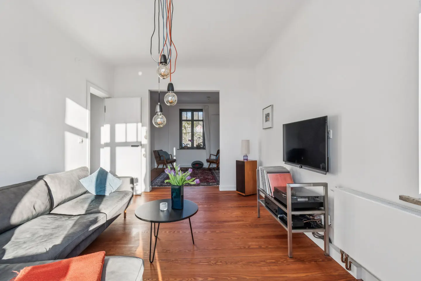 Bright living room with gray sofa, black coffee table with tulips, and a TV on the white wall. Hardwood floors and pendant lights add warmth.