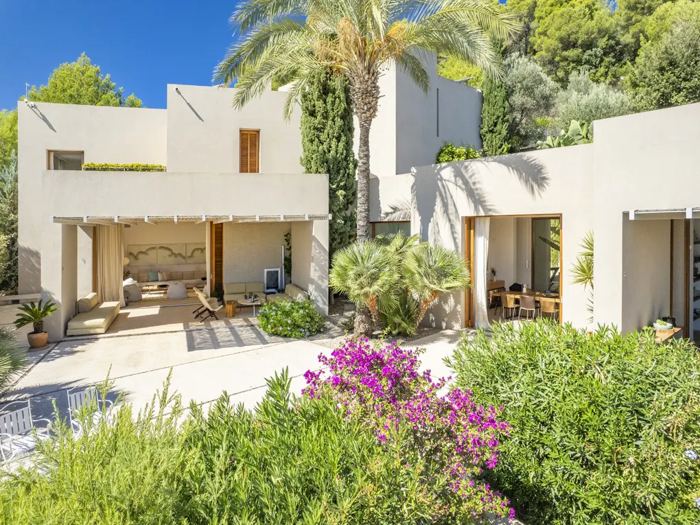 Exterior of a modern, off-white home with a palm tree, lush greenery, and purple flowers in the foreground. Outdoor seating areas are visible.