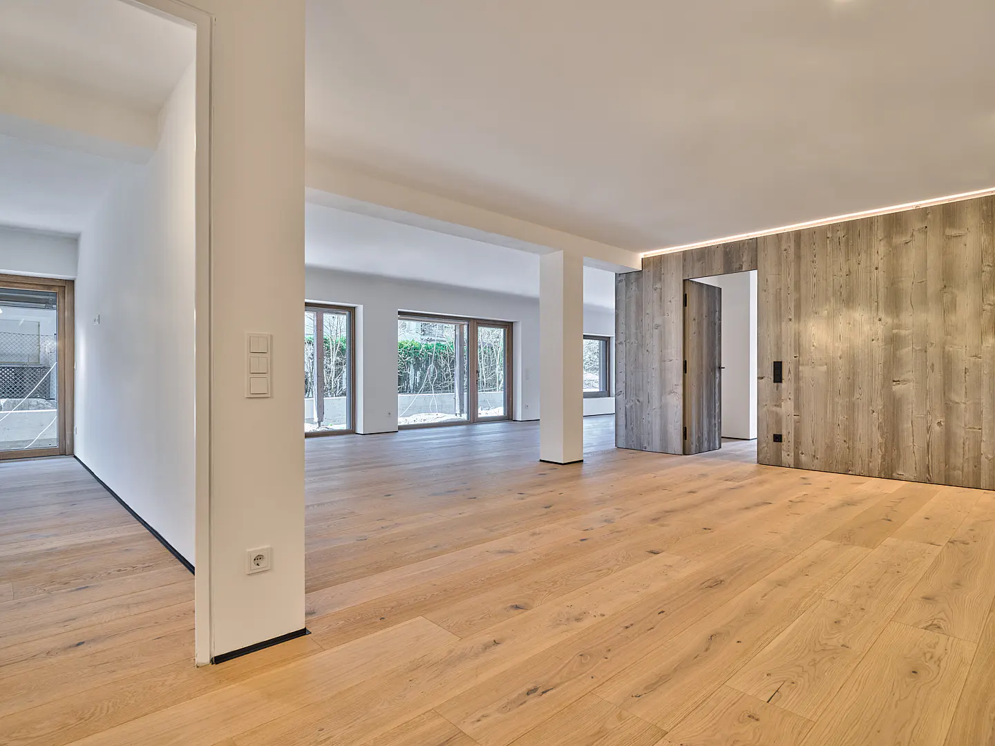 Bright, empty room with light wood floors, white walls, and large windows. A wood-paneled wall with a doorway adds texture.