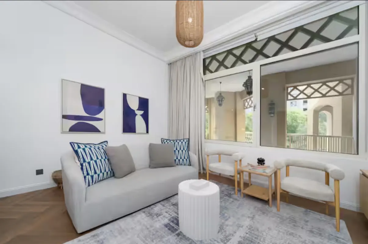 Bright living room with white sofa, blue pillows, and abstract art. Large windows offer a view of a balcony.