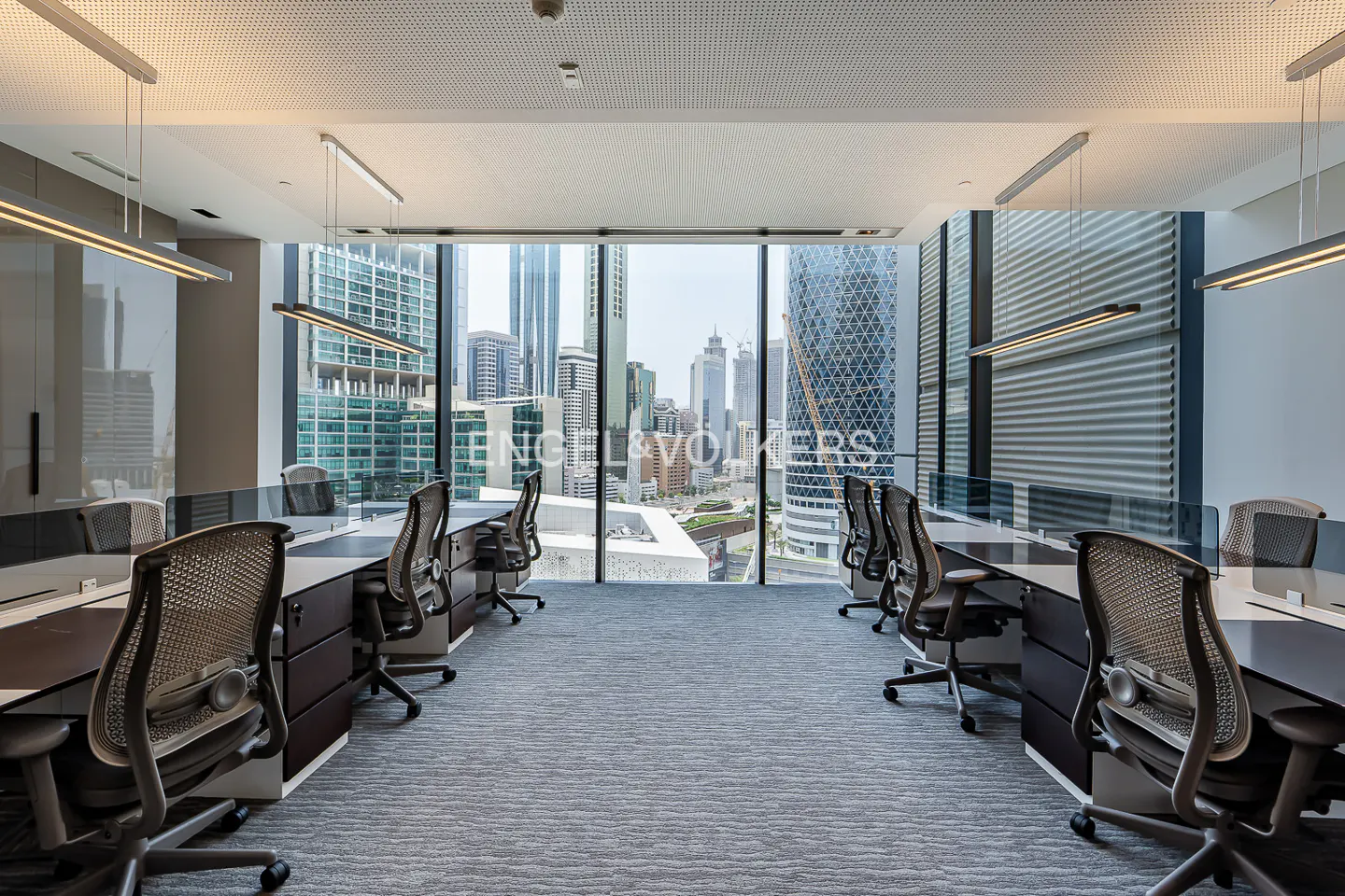 Office space with desks and chairs facing a large window with a city view. Gray carpet and modern lighting.