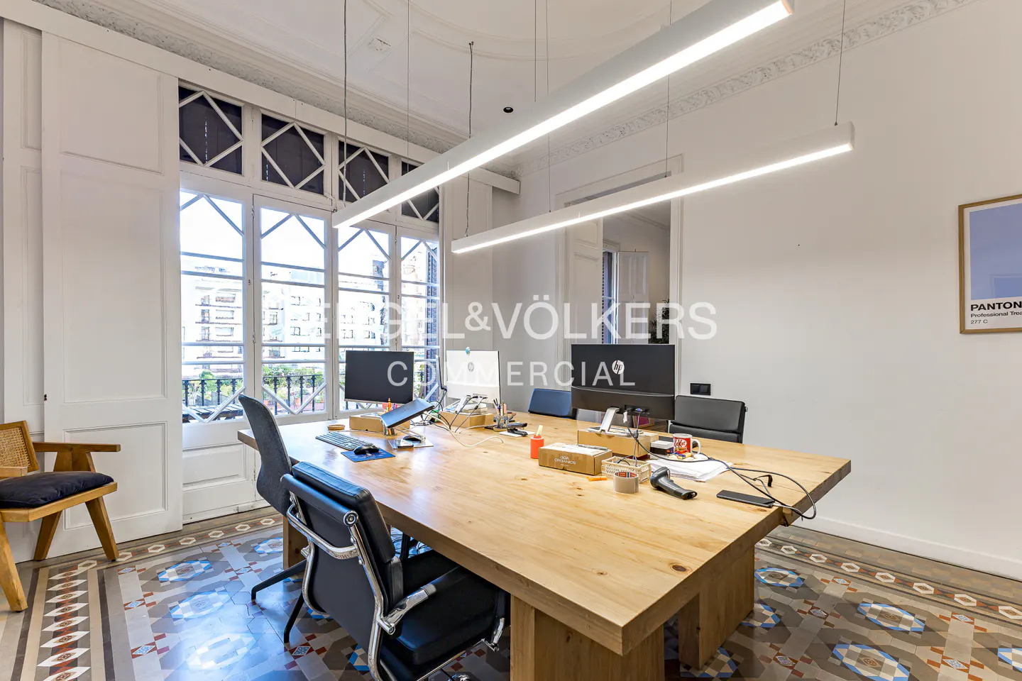 Bright office space with a large wooden table, black chairs, and computers. Large windows offer a city view. Ornate tile floor and modern lighting.