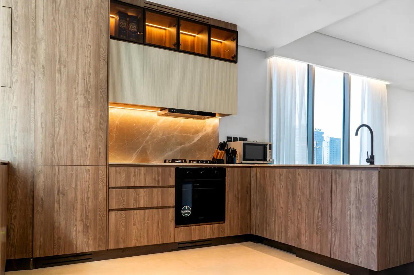 Modern kitchen with wood cabinets, marble backsplash, black oven, and stainless steel microwave. A window with sheer curtains is visible.