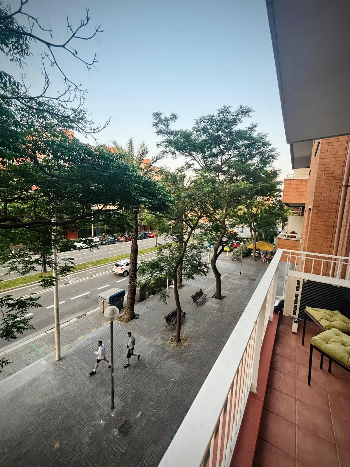 View from a balcony with white railings overlooking a tree-lined street with two people running on the sidewalk.