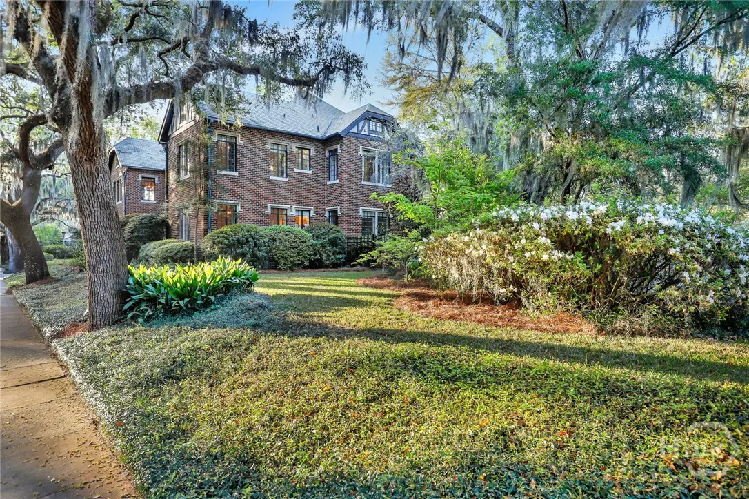 A two-story brick house with a gray roof, surrounded by green bushes and trees.