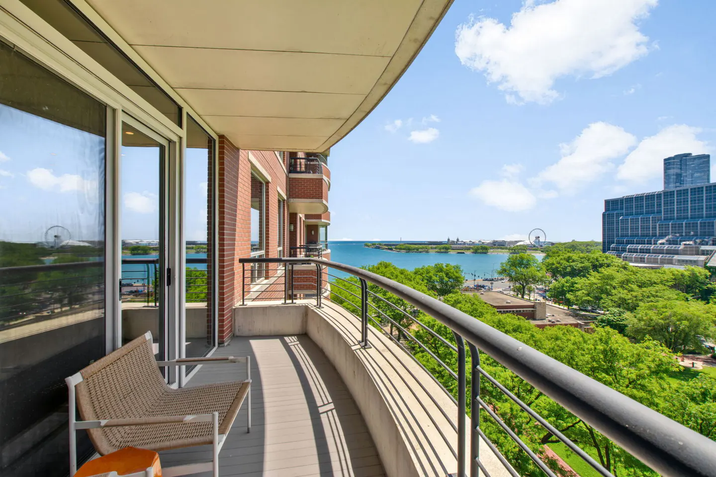 Curved balcony with a wicker chair overlooking a lush park, blue water, and city skyline under a bright sunny sky.