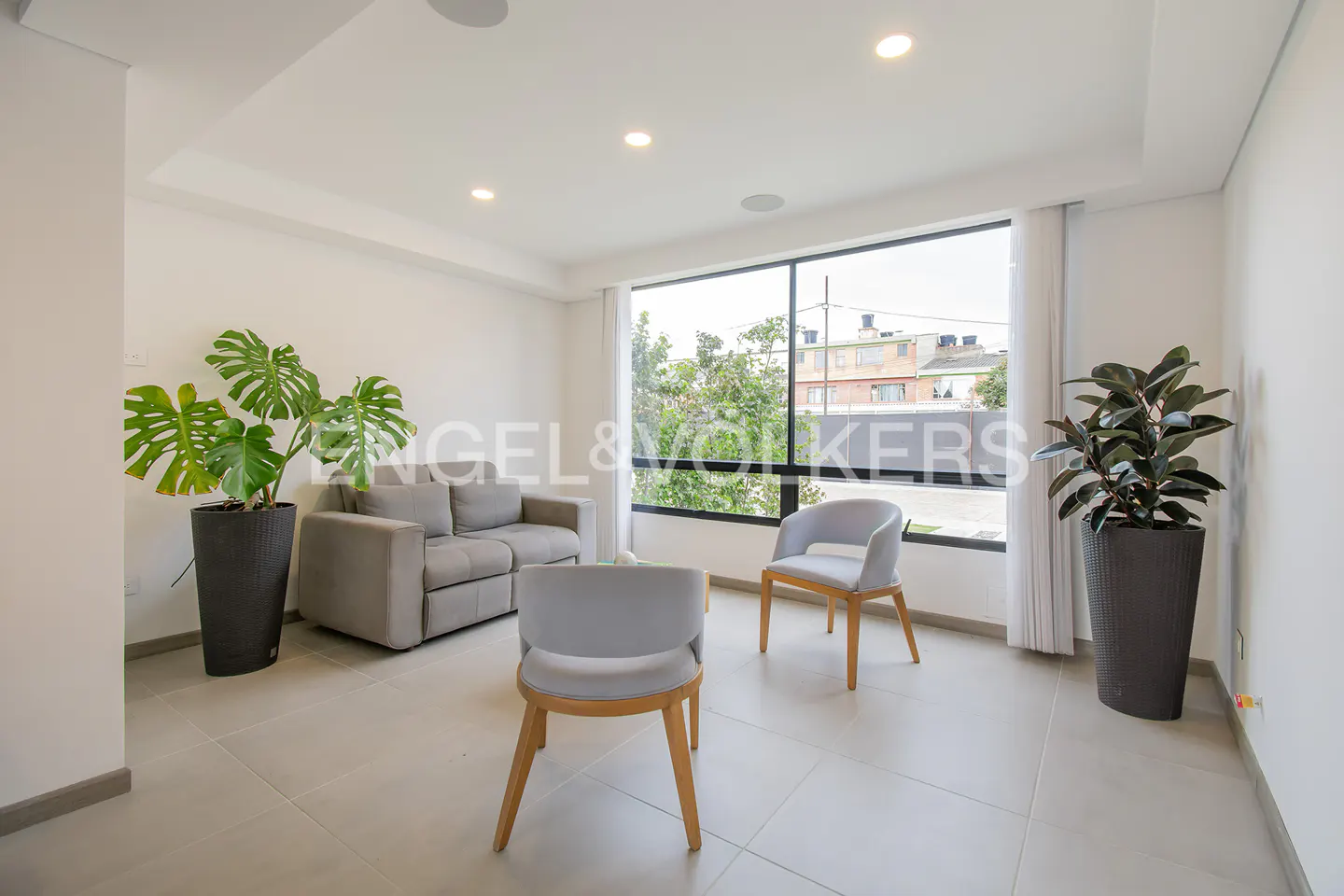 Bright living room with gray sofa, two light gray chairs, and potted plants by a large window.