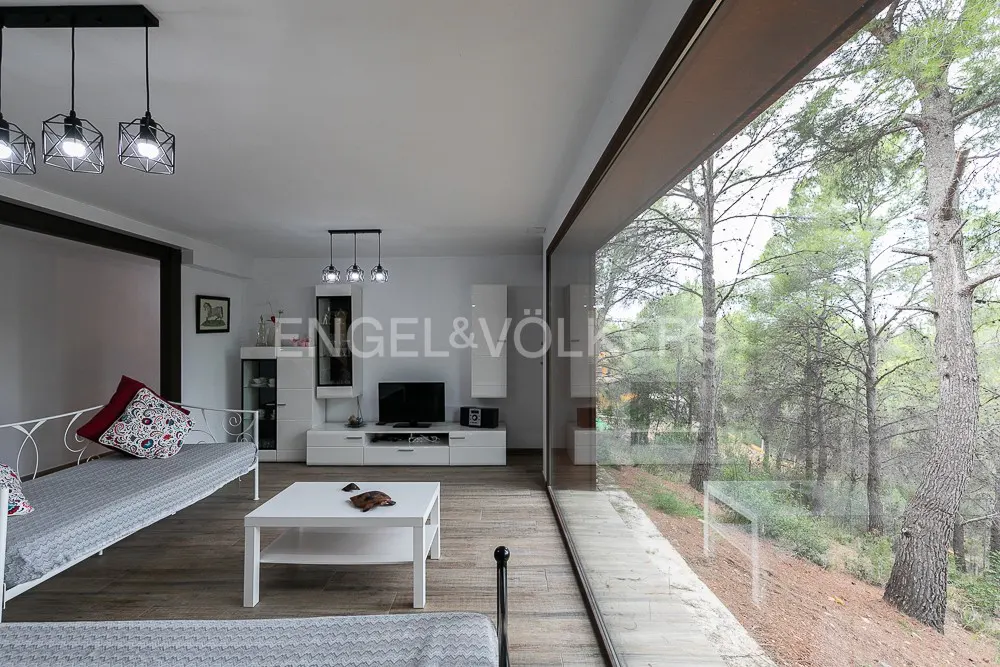 Living room with white furniture, wood floors, and a large window overlooking a forest. Black pendant lights hang from the ceiling.
