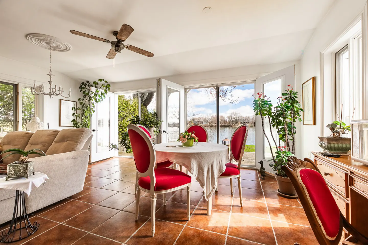 Bright sunroom with terracotta tile, white walls, and a view of the lake. A round table with red chairs sits in the center.