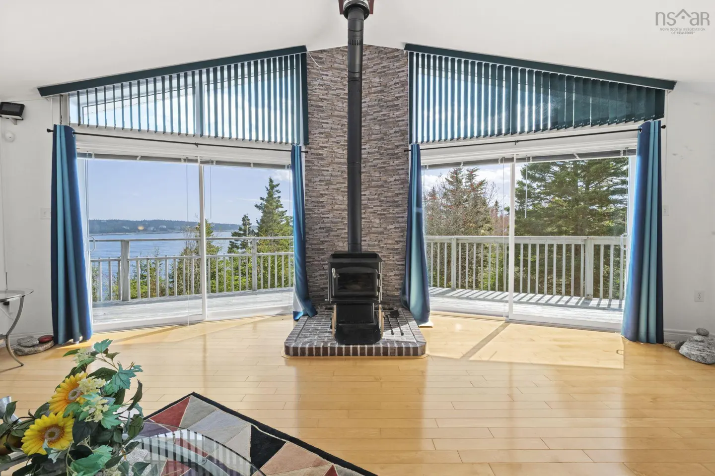 Living room with a central stone fireplace, wood stove, and large windows overlooking a deck and water view.