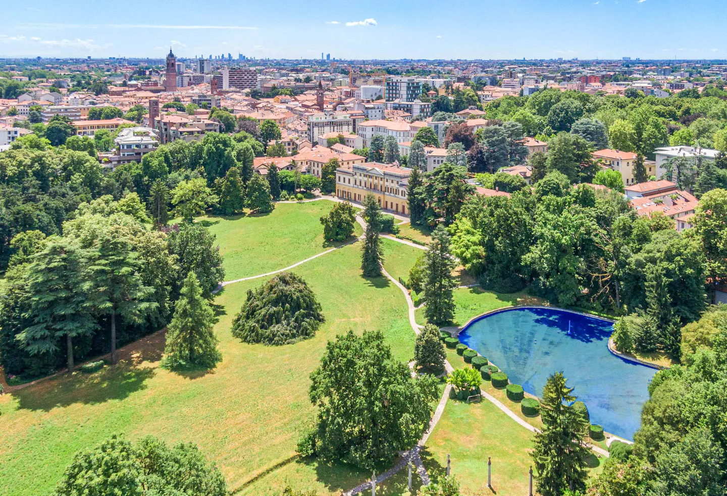 Aerial view of a yellow villa with a blue pond, green lawn, and trees in a city park on a sunny day.