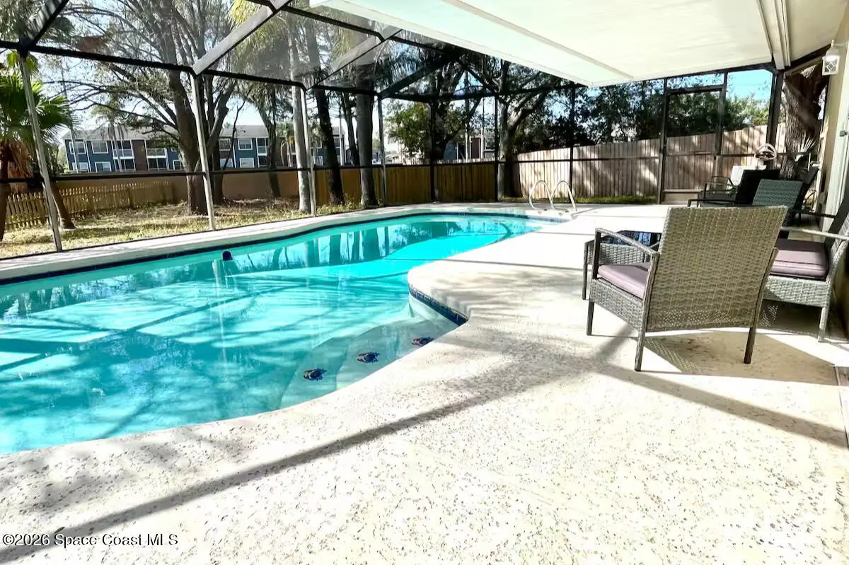 Screened-in pool with turquoise water, patio furniture, and a view of trees and a fence.
