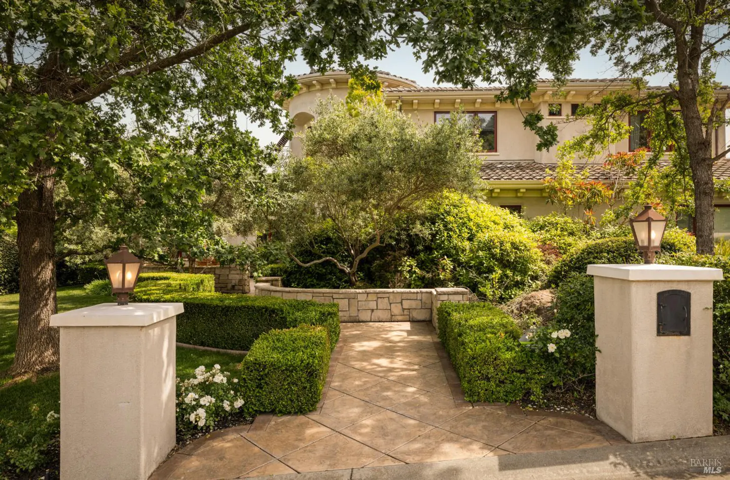 Landscaped walkway to a beige house. Stone pillars with lamps flank the path lined with trimmed hedges and lush greenery.