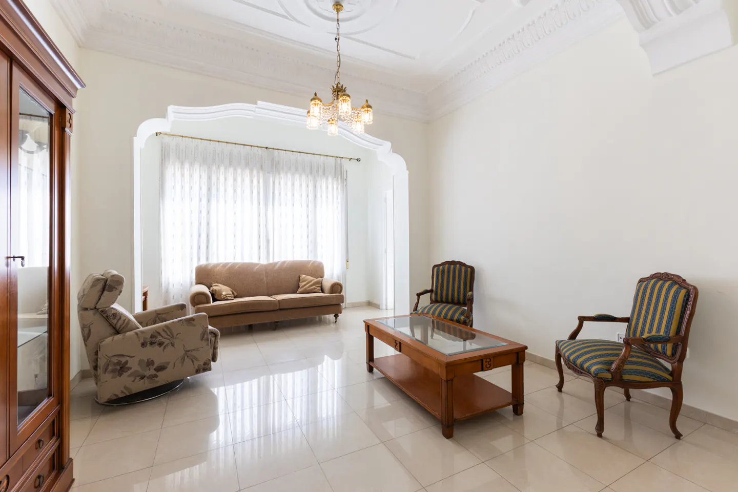 Bright living room with beige sofa, striped chairs, and wooden coffee table on a tiled floor. A chandelier hangs from the ornate ceiling.