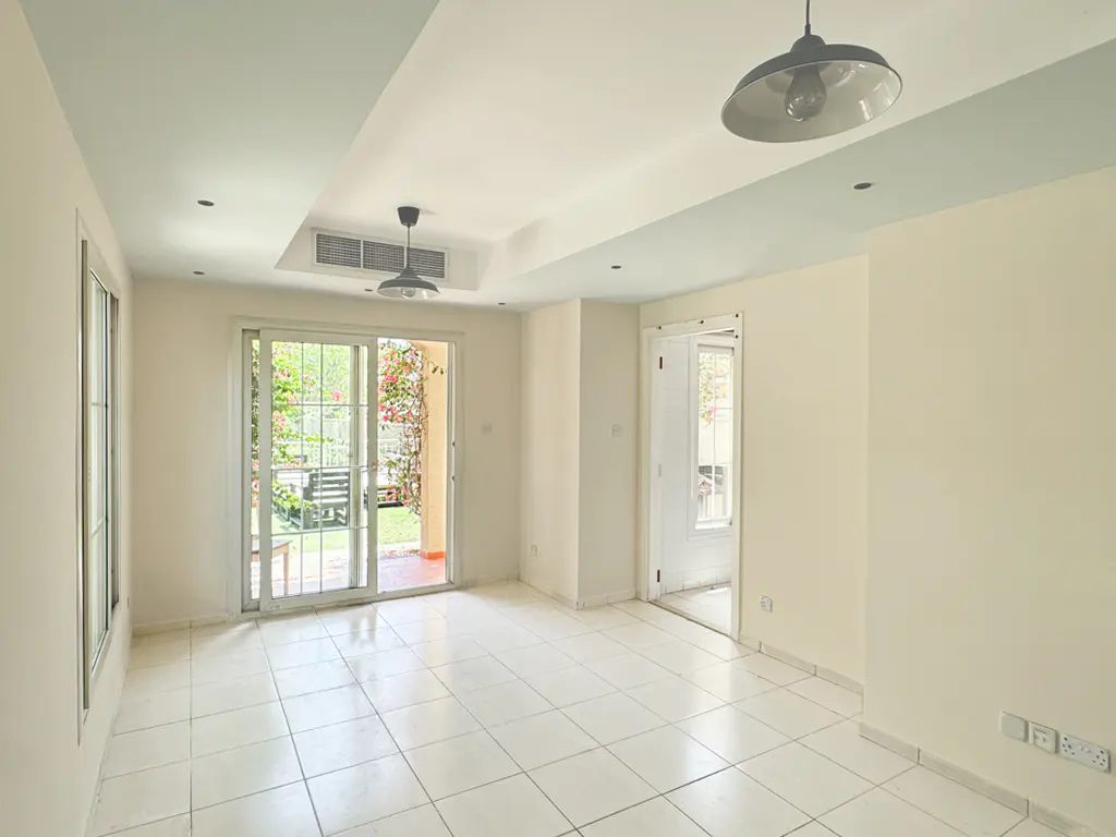 Bright, empty room with white tile floor, cream walls, and sliding glass doors to a garden. Two pendant lights hang from the ceiling.
