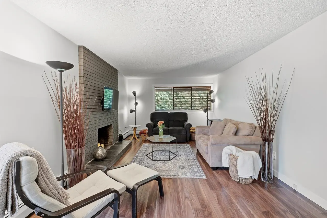 Living room with hardwood floors, brick fireplace, TV, black and beige sofas, and a patterned rug. Tall vases with branches add decor.