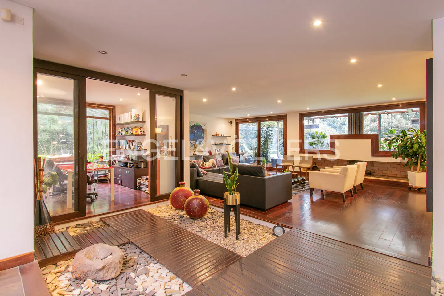 Open-concept living room with wood floors, a gray sofa, and two cream-colored armchairs. A zen garden is in the foreground.