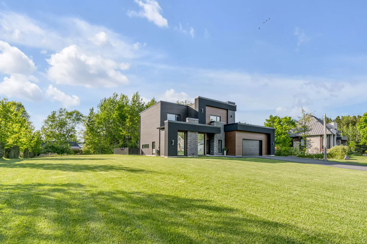 Modern two-story home with black and wood siding, stone accents, and a large green lawn under a bright blue sky.