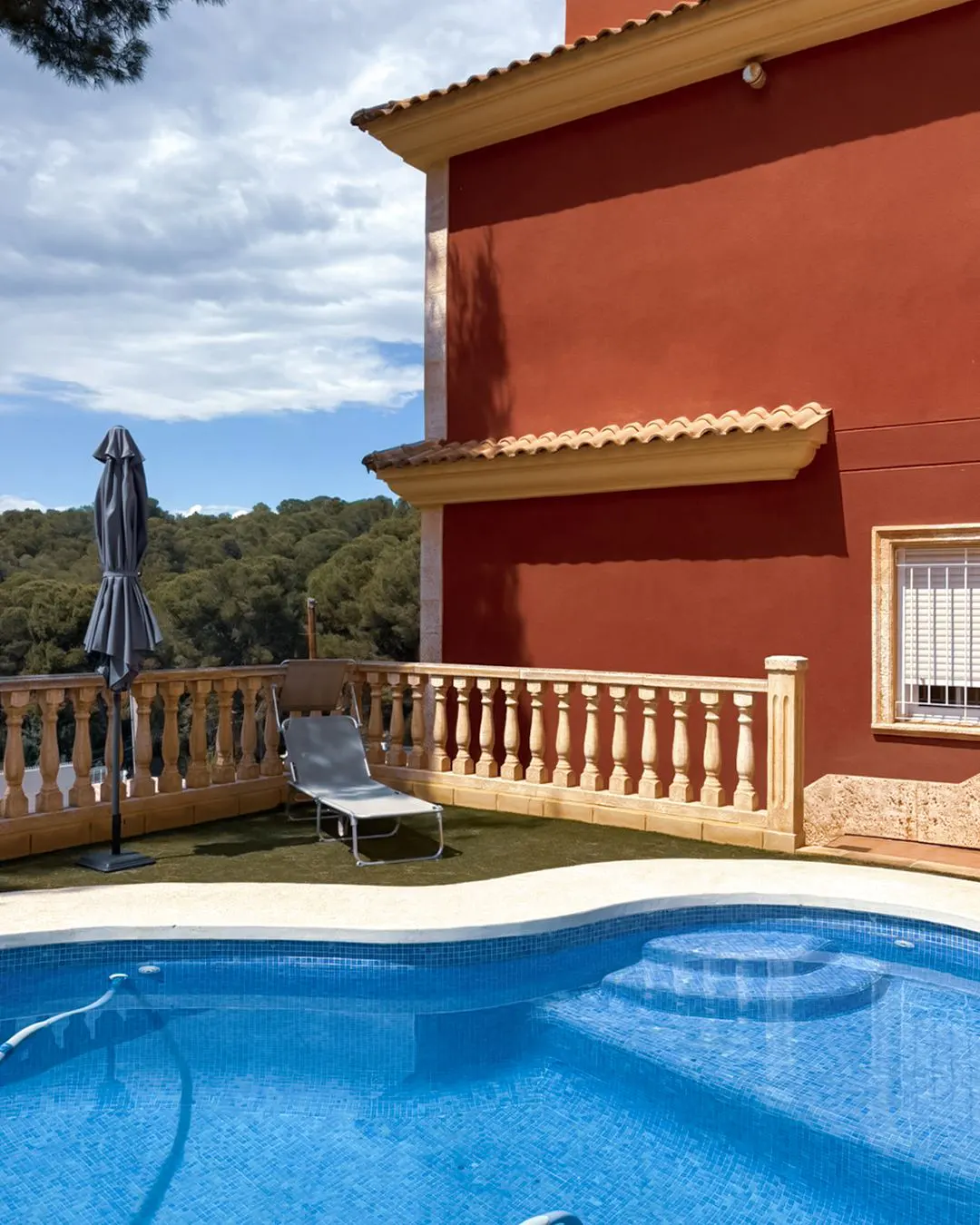 Outdoor pool with blue tiles, lounge chair, and closed umbrella. Red house with tan trim and balustrade. Forest in the background.