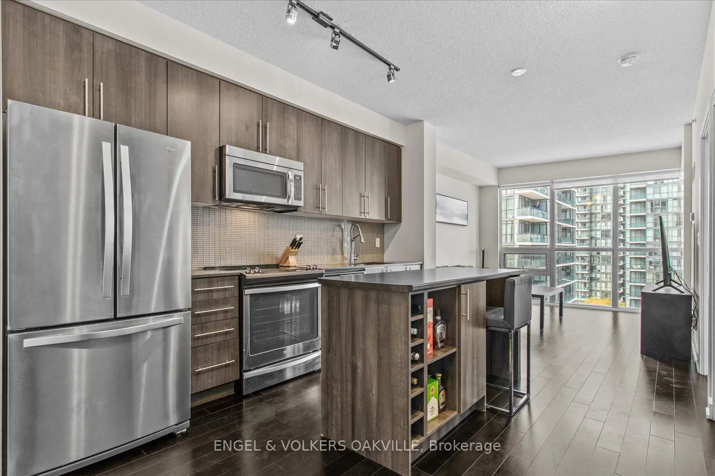 Modern kitchen with stainless steel appliances, wood cabinets, and dark wood floors. Island with seating and wine rack. Large windows with city views.