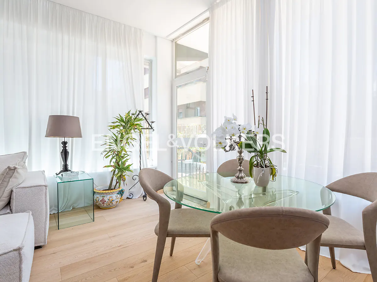 Bright dining area with a glass table, beige chairs, and white orchids. Sheer white curtains filter light from a window. A lamp and plant sit nearby.