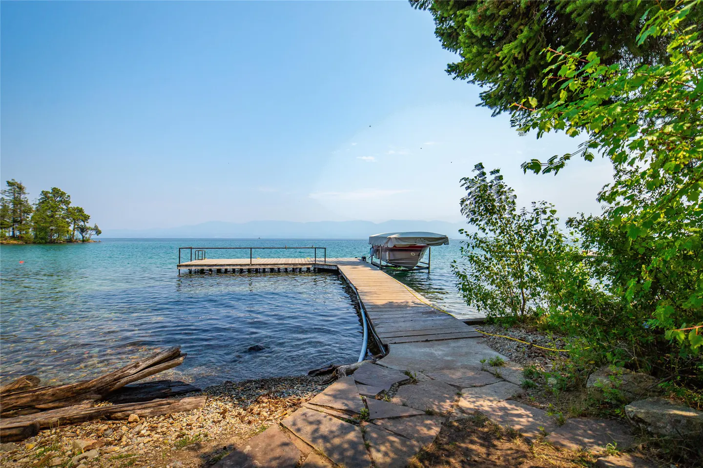 Scenic view of a lake with a wooden dock and boat lift under a clear blue sky. Lush greenery frames the right side of the image.