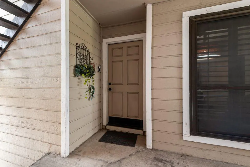 Exterior view of a beige apartment entrance with a brown door, black doormat, and a decorative plant hanger. A window with blinds is visible to the right.