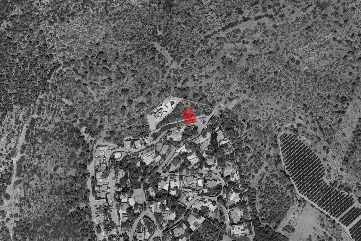 Aerial view of a property in a wooded area, outlined in red, near a residential area in grayscale.