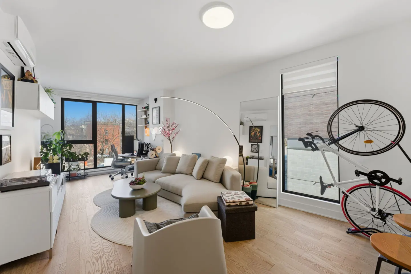 Bright living room with a beige sectional sofa, round coffee table, and a bicycle rack near the window. Hardwood floors and white walls.