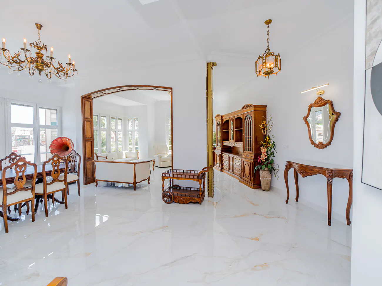 Bright dining and living area with white marble floors, ornate chandeliers, wooden furniture, and a vintage gramophone.