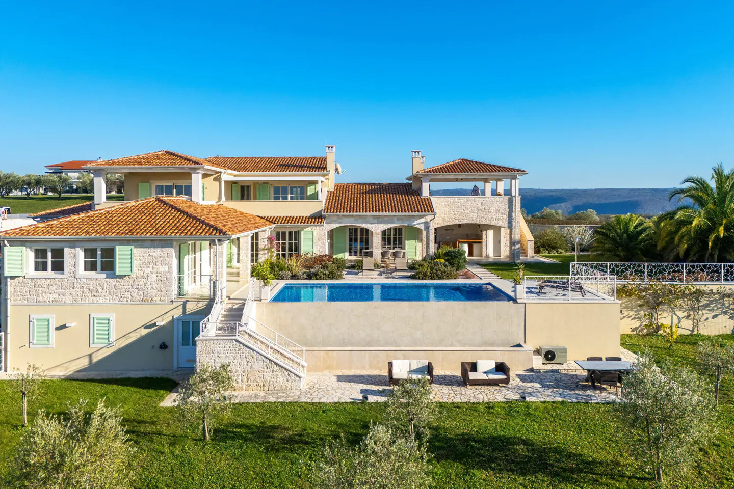 Luxury villa with a pool. The house has a red tile roof, light yellow walls, and green shutters. Blue sky background.