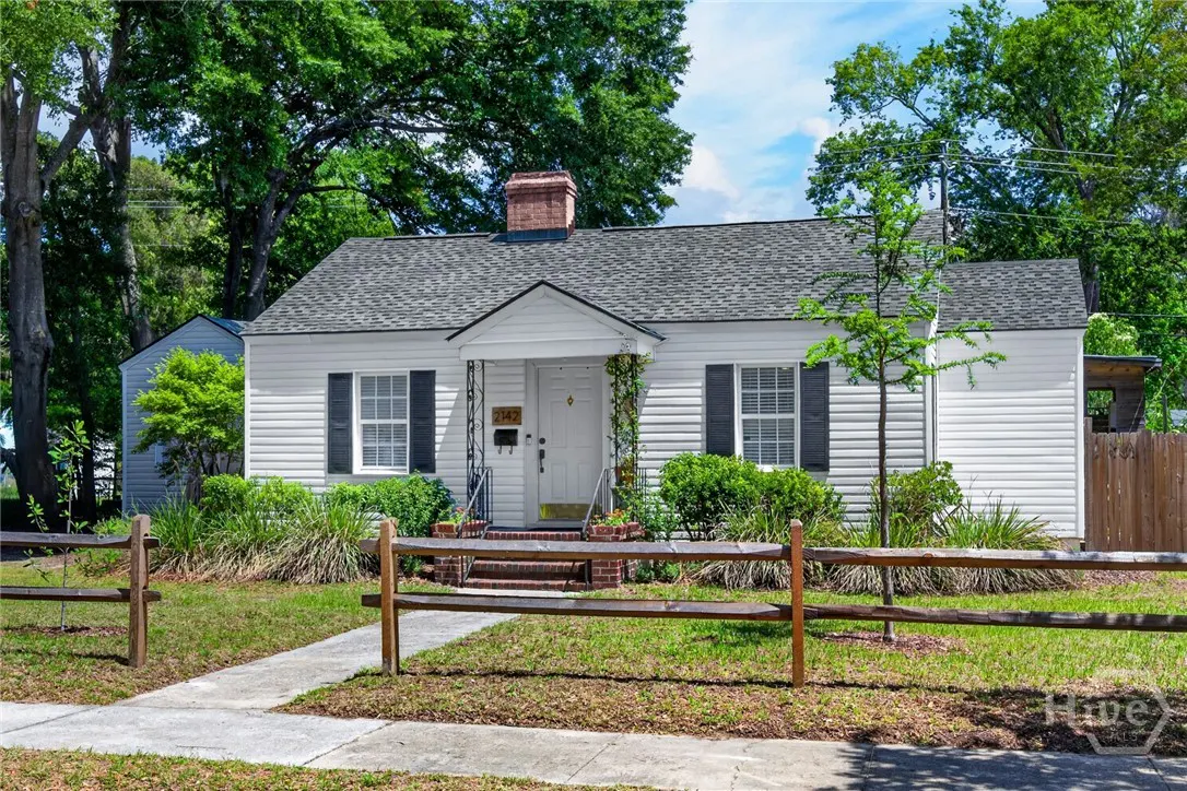 A white, one-story house with black shutters, a gray roof, and a brick chimney, surrounded by green trees and a wooden fence.