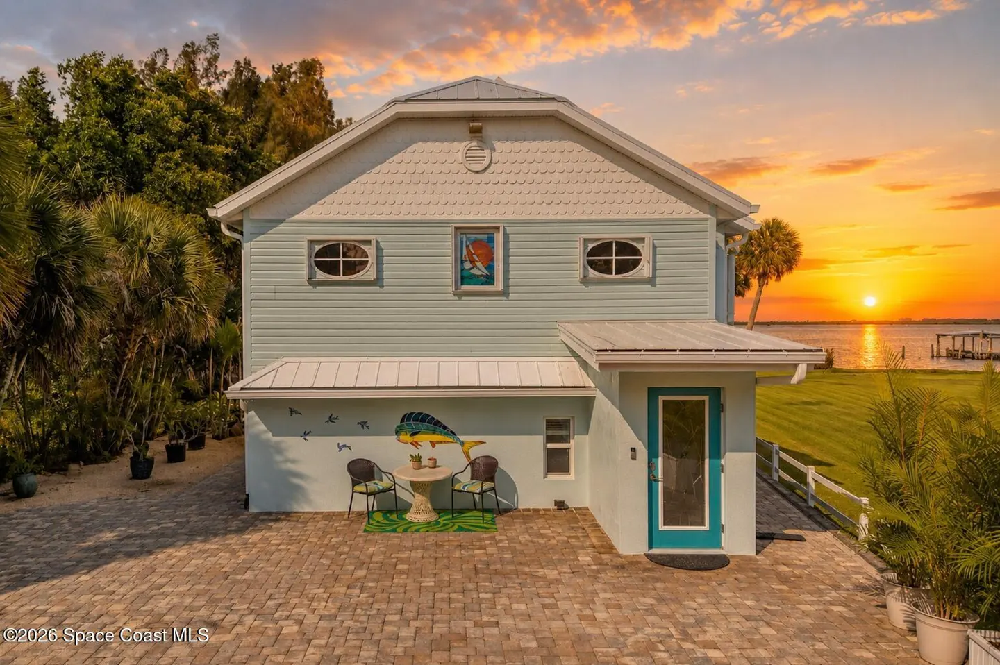 Two-story light blue house with white trim, a mural of a fish, and a sunset over the water in the background.