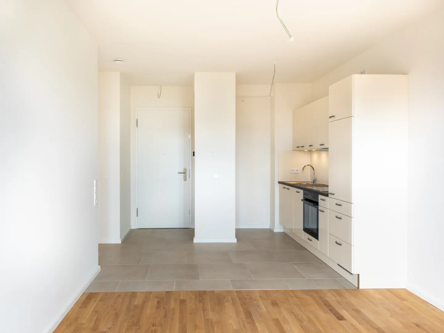 Bright, empty apartment with white walls, tile and wood floors, and a small white kitchen. A white door is visible in the background.