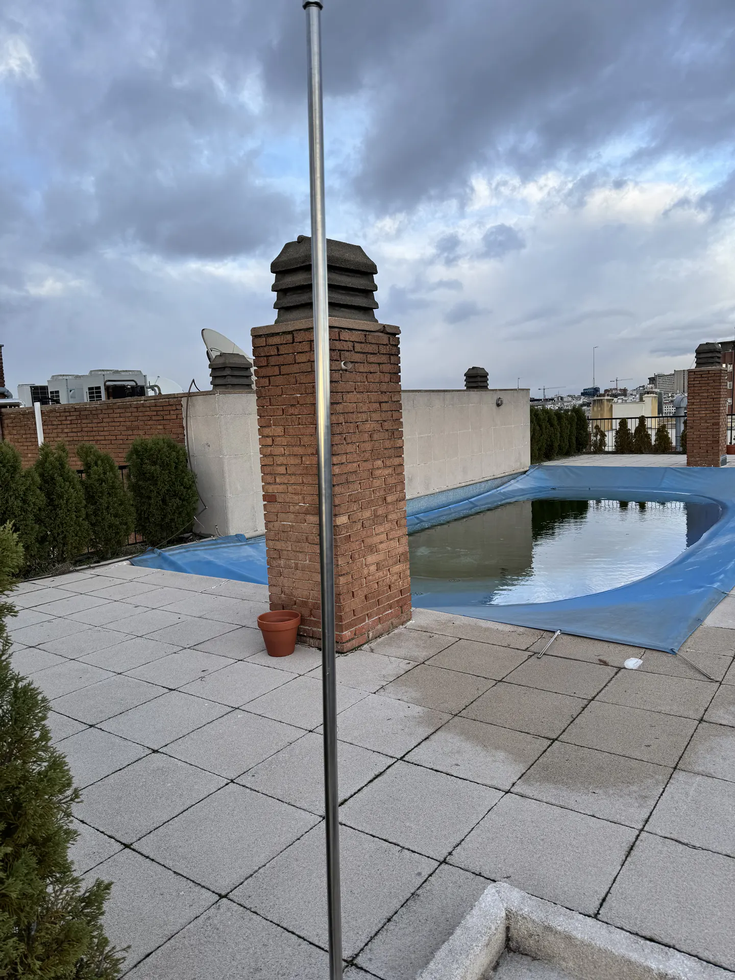 Rooftop pool with a blue cover, brick chimney, and gray tiled deck under a cloudy sky.
