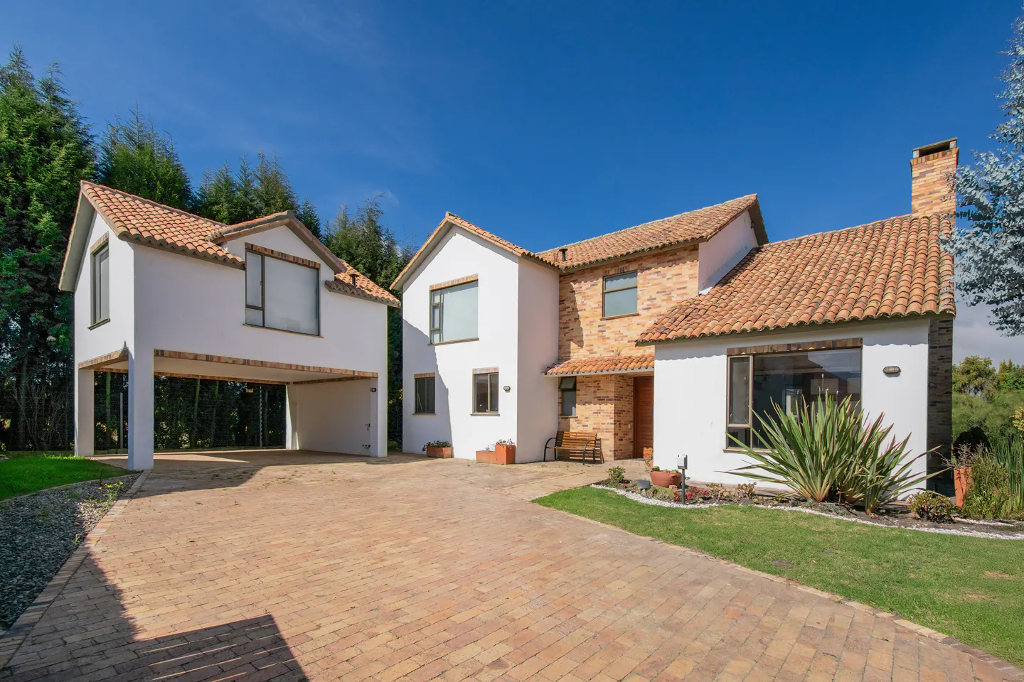 Exterior of a two-story house with white walls, brick accents, and a terracotta tile roof. A brick driveway leads to a covered carport.