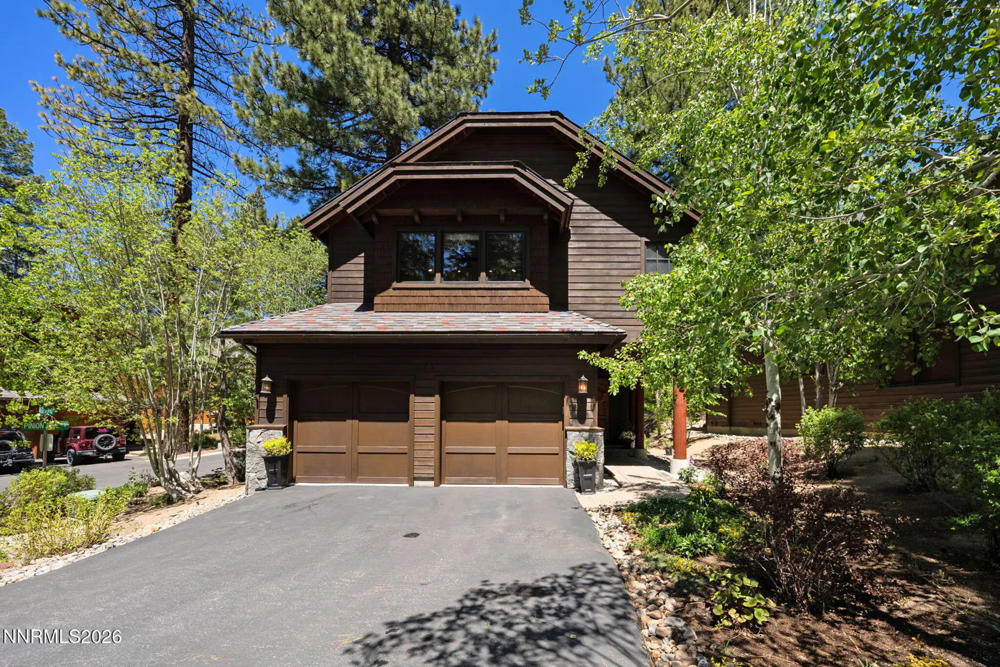 Two-story brown house with a two-car garage and a dark asphalt driveway. Green trees surround the house under a blue sky.