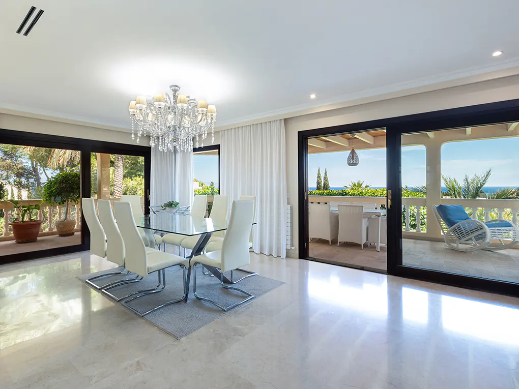 Bright dining room with glass table, white chairs, and crystal chandelier. Sliding doors open to a balcony with ocean views.