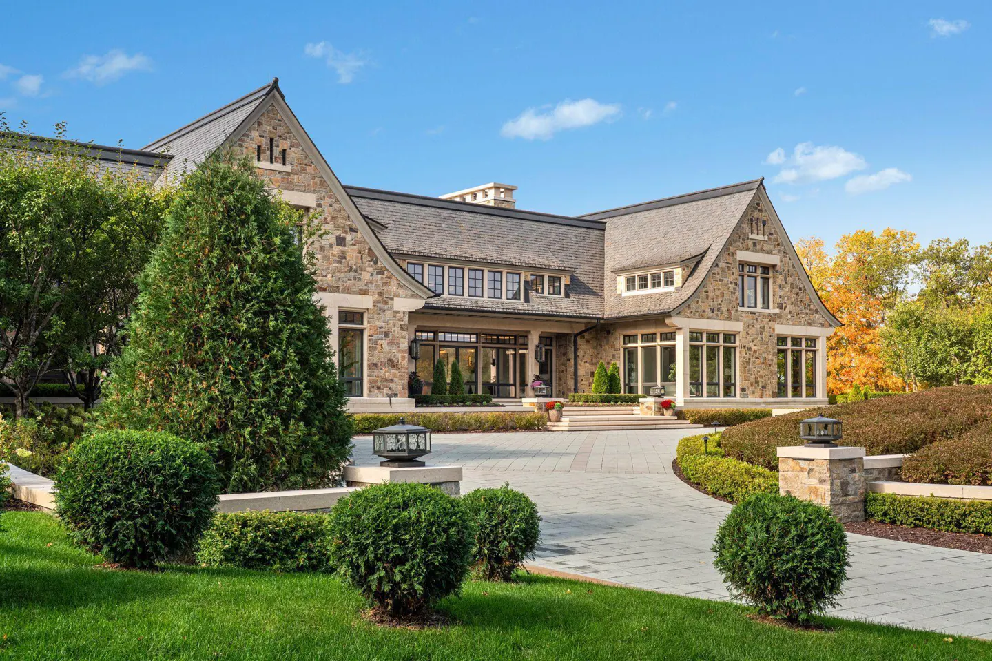 Stone mansion with a gray roof, manicured lawn, and a paved driveway under a blue sky.