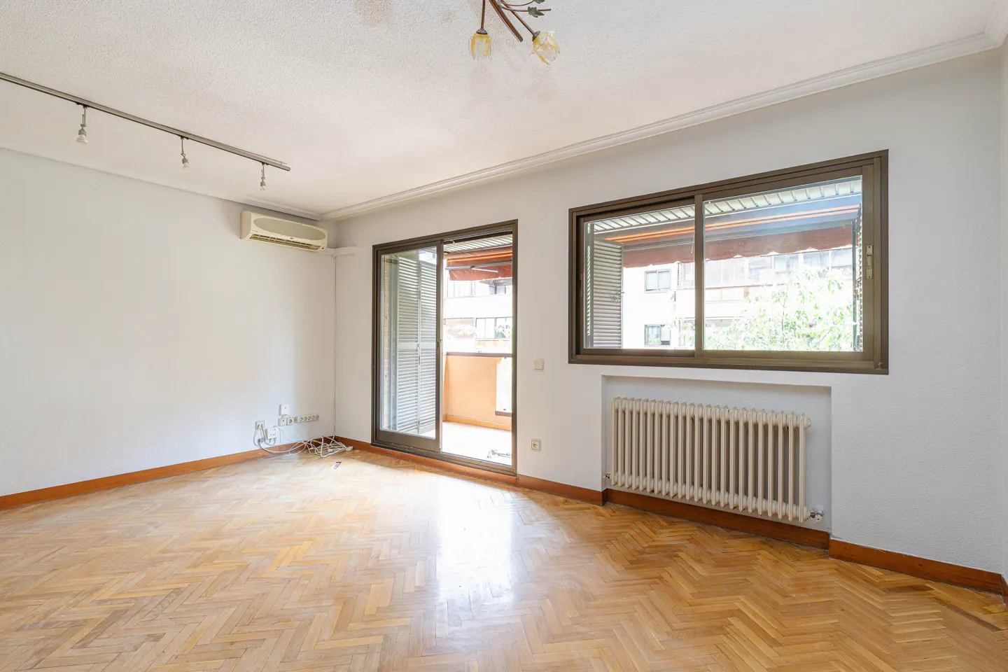 Bright, empty room with herringbone wood floors, white walls, and a sliding glass door to a balcony. A radiator sits under a window.