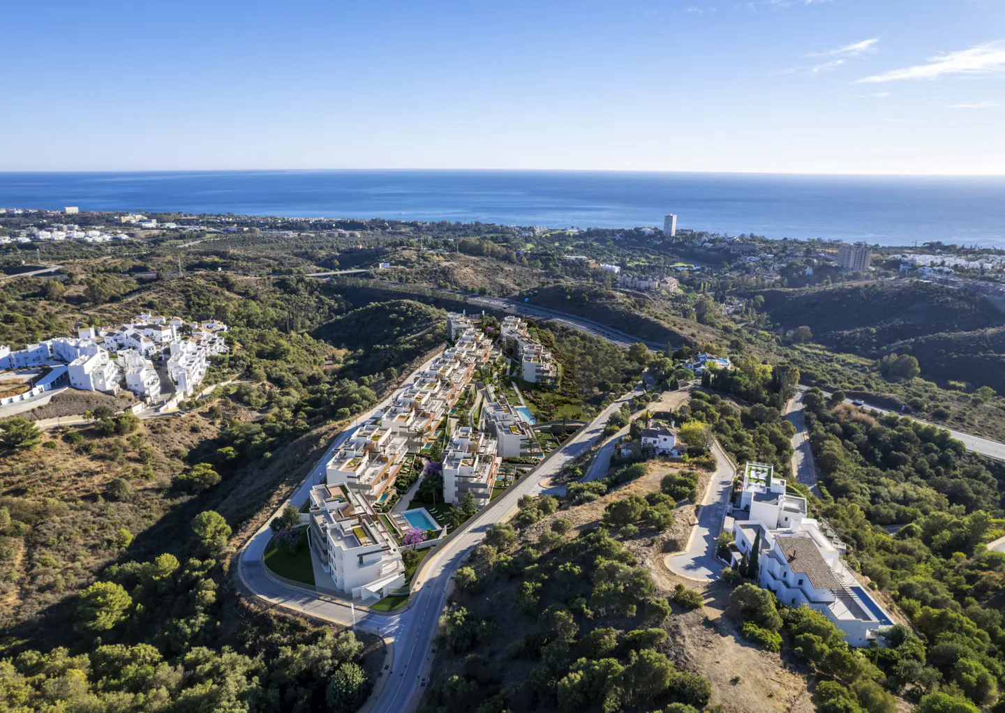 Aerial view of white modern villas with pools nestled in green hills, overlooking the blue ocean under a clear sky.