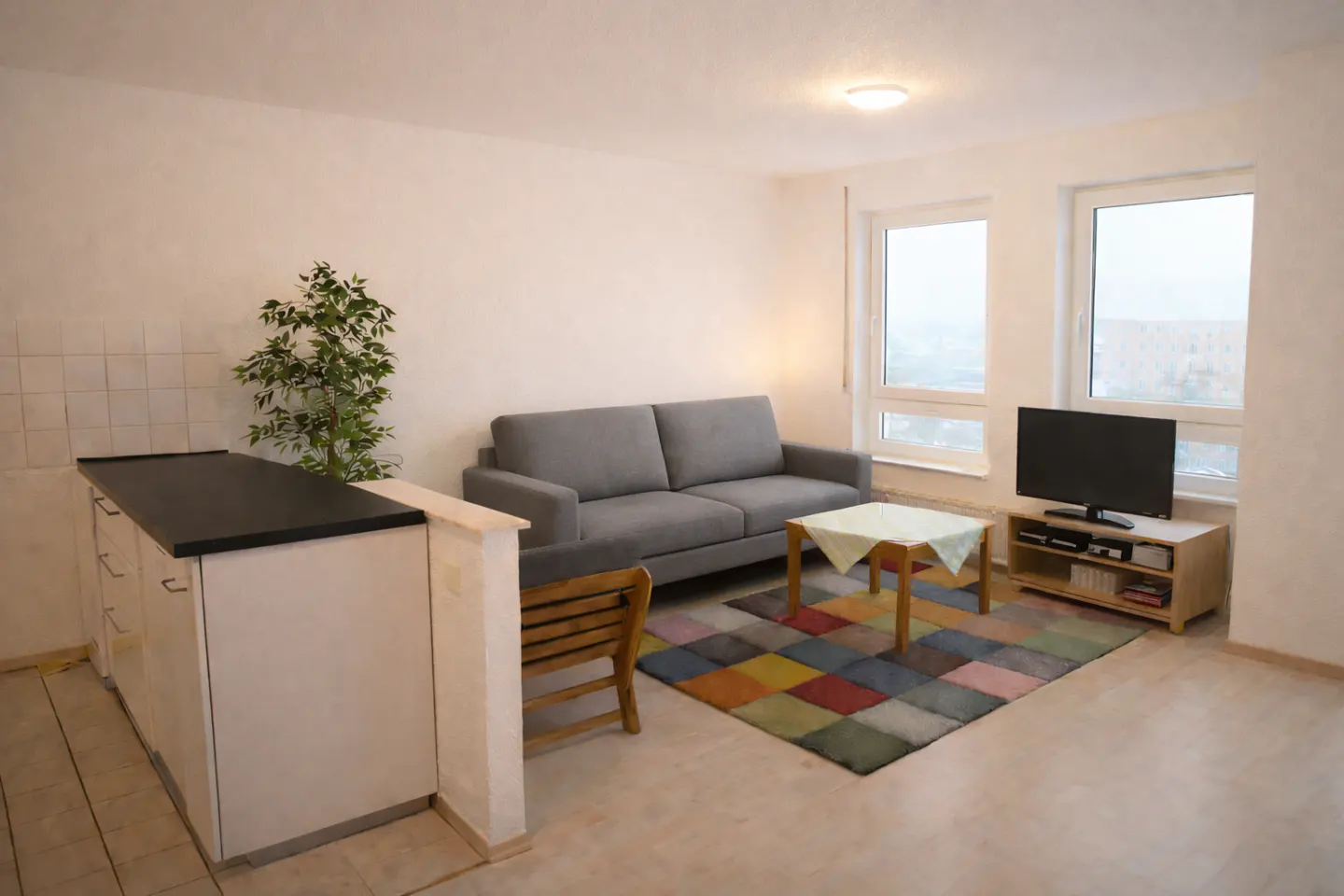 A bright living room with a gray sofa, TV, and colorful rug. Windows overlook a cityscape. A kitchen counter is on the left.