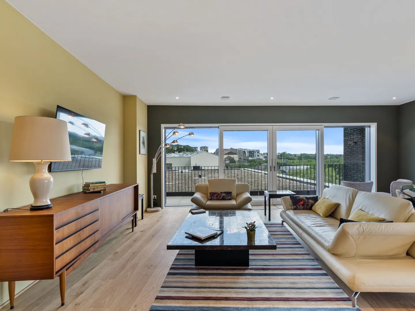 Living room with a tan sofa, chair, and wood cabinet. A striped rug sits on the wood floor. Balcony doors open to a city view.